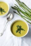 Two bowls of asparagus soup topped with asparagus next to a spoon and fresh asparagus
