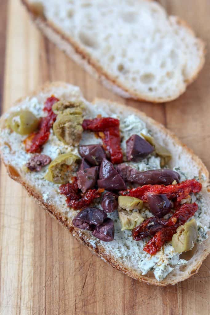 A slice of bread topped with goat cheese, olives and sun-dried tomatoes sitting on a cutting board