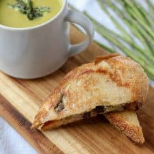 A mug of asparagus soup next to a grilled cheese sandwich on a cutting board