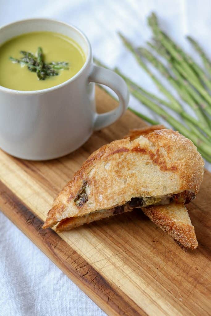 A mug of asparagus soup next to a grilled cheese sandwich on a cutting board