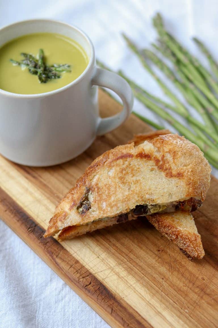 A mug of asparagus soup next to a grilled cheese sandwich on a cutting board