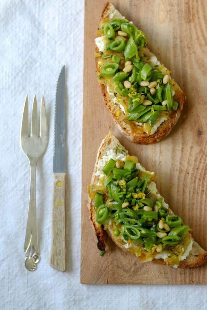 Open sandwiches topped with snap peas and leeks on a wooden cutting board next to a knife and fork