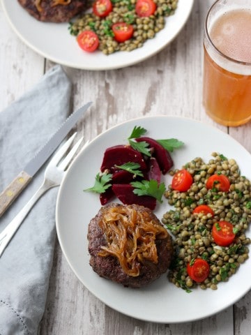 A hamburger, pickled beets and lentil salad on a plate next to a fork, knife, napkin and glass of beer