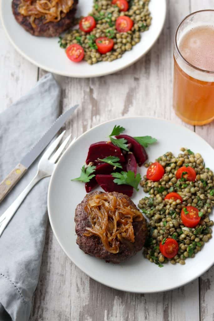 A hamburger, pickled beets and lentil salad on a plate next to a fork, knife, napkin and glass of beer