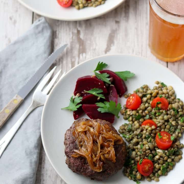 A hamburger, pickled beets and lentil salad on a plate next to a fork, knife, napkin and glass of beer