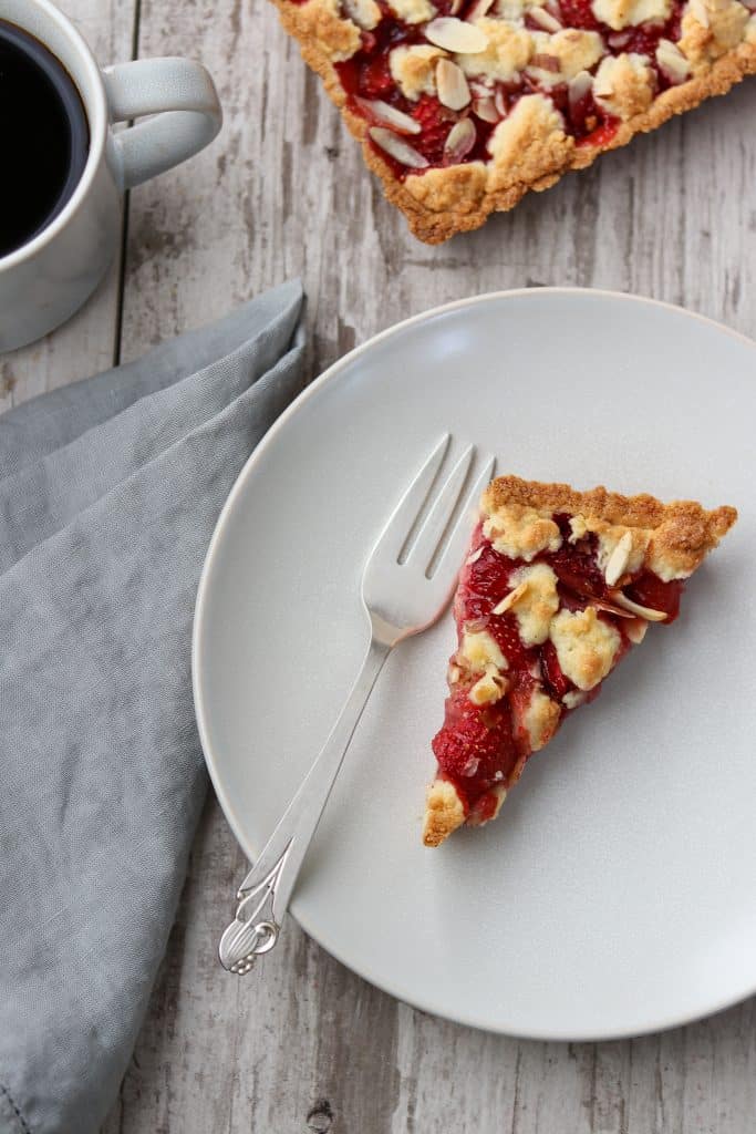 A slice of strawberry almond tart on a plate with a fork, napkin and cup of coffee