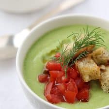 A bowl of cucumber soup topped with tomatoes, croutons and dill next to a spoon