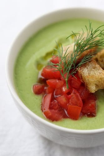 A close up of a bowl of cucumber soup with chopped tomatoes, croutons and dill