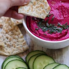 A bowl of beet hummus next to flatbread and cucumbers