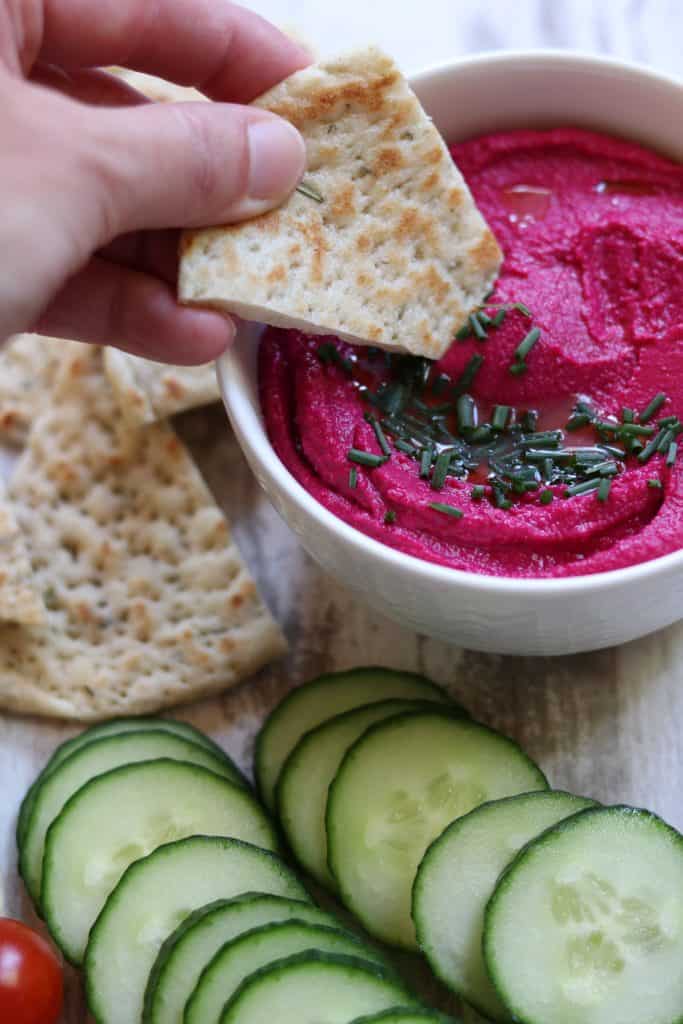 A bowl of beet hummus next to flatbread and cucumbers