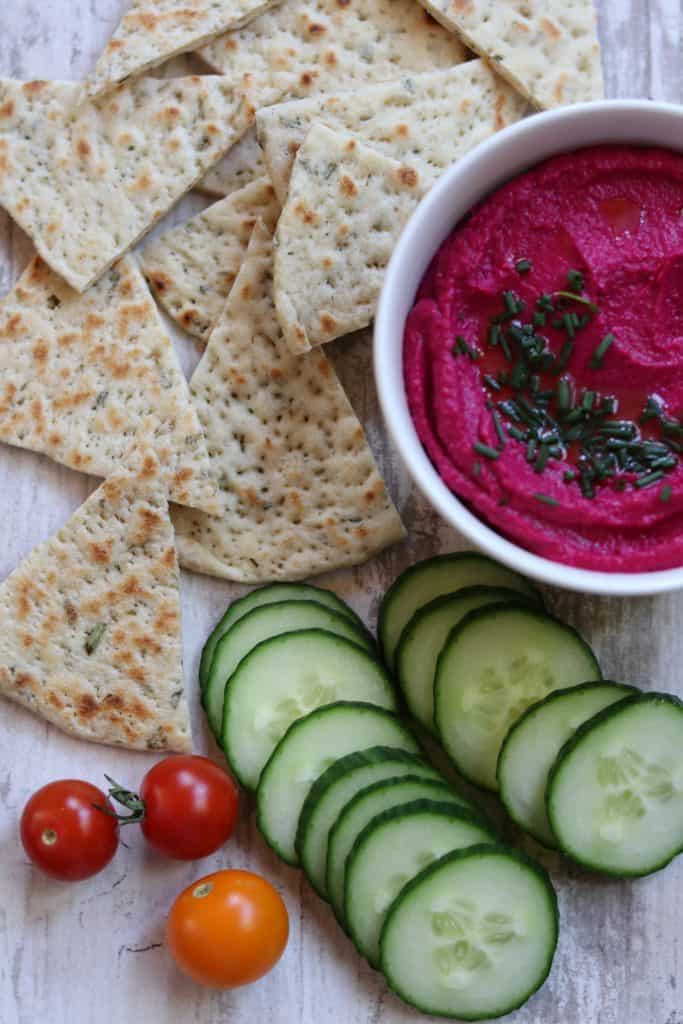 A bowl of beet hummus next to flatbread, cucumbers and tomatoes