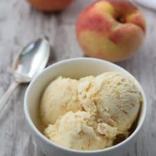 A bowl of roasted peach ginger ice cream next to a spoon and fresh peaches