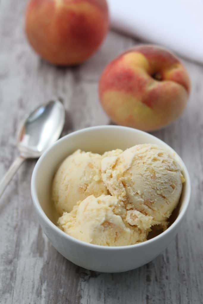 A bowl of roasted peach ginger ice cream next to a spoon and fresh peaches