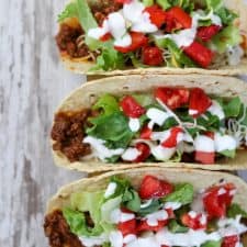 Three tacos with toppings on a wooden surface