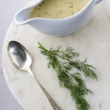 Creamy mustard dill sauce in a gravy boat with a spoon and fresh dill on a marble surface