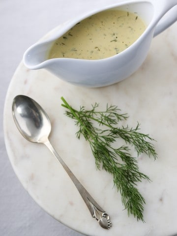Creamy mustard dill sauce in a gravy boat with a spoon and fresh dill on a marble surface