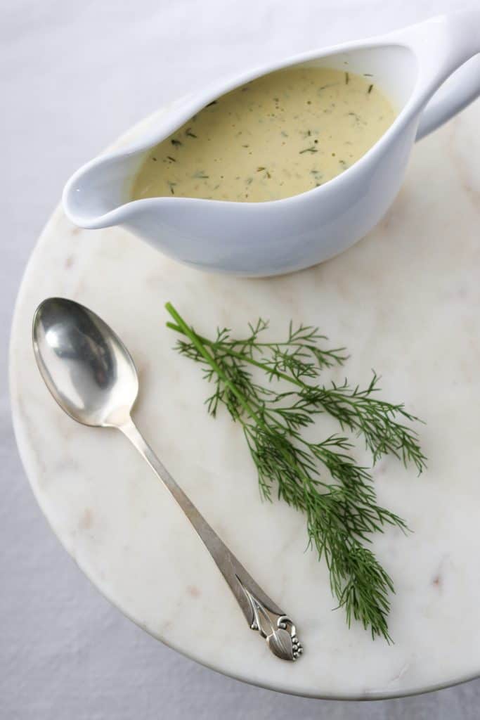 Creamy mustard dill sauce in a gravy boat with a spoon and fresh dill on a marble surface