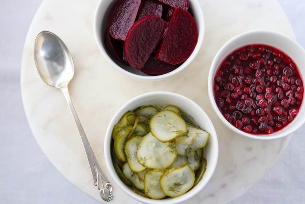 Bowls of roasted beets, pickles and lingonberries with a spoon on a marble surface