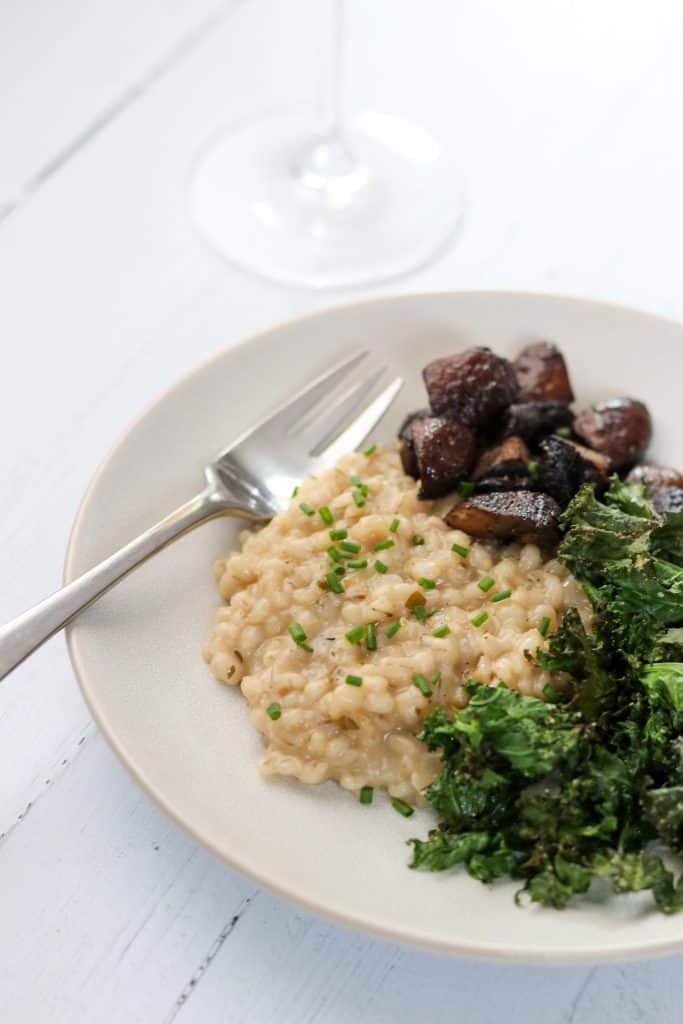 A plate of barley risotto, roasted mushrooms and kale on a wood surface with a wine glass