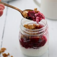 A close up of yogurt topped with raspberry, roasted beets and granola in a jar with a spoon