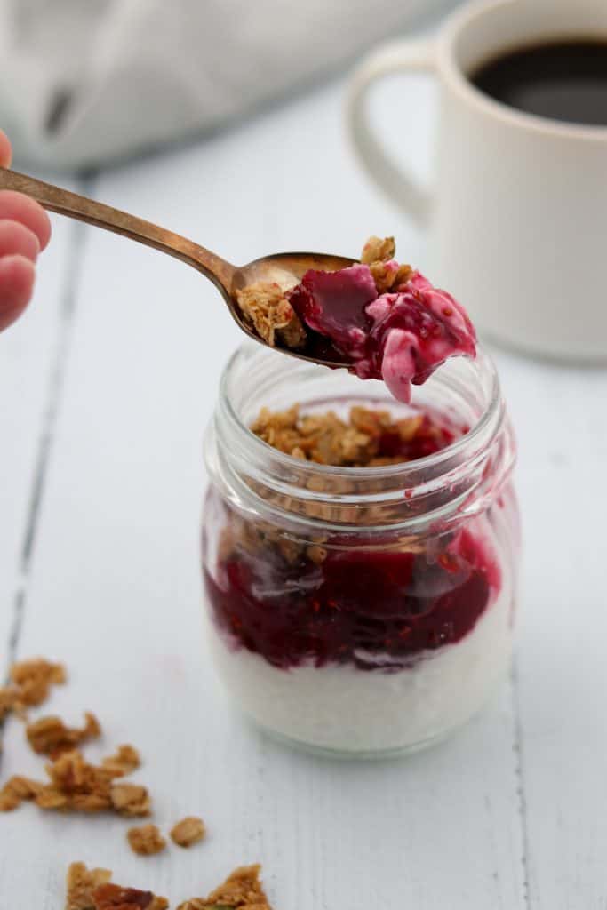 A close up of yogurt topped with raspberry, roasted beets and granola in a jar with a spoon