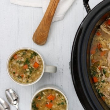 Chicken soup in a crockpot with two mugs of soup, spoons and a wooden ladle