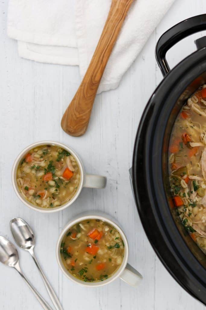 Chicken soup in a crockpot next to two mugs of soup, spoons and a ladle