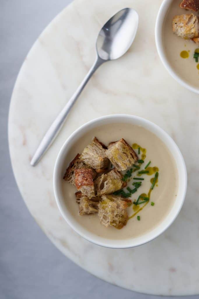 A bowl of cauliflower soup with croutons, chives and olive oil on a plate with a spoon