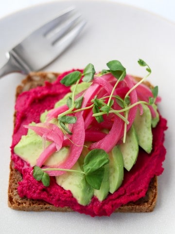 An open sandwich with beet hummus, avocado slices and pickled onions on a plate with a fork