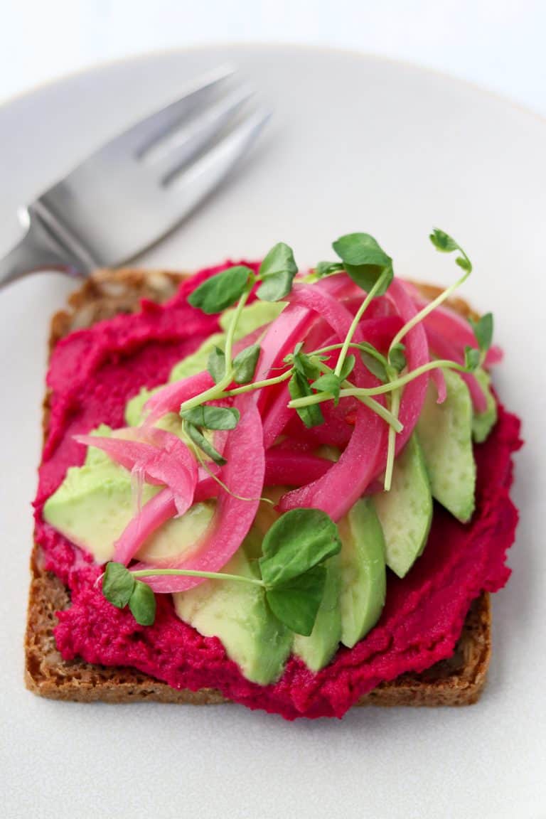 An open sandwich with beet hummus, avocado slices and pickled onions on a plate with a fork