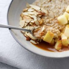 Oatmeal in bowl topped with apple, sliced almonds and caramel sauce with a spoon