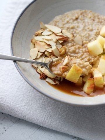 Oatmeal in bowl topped with apple, sliced almonds and caramel sauce with a spoon