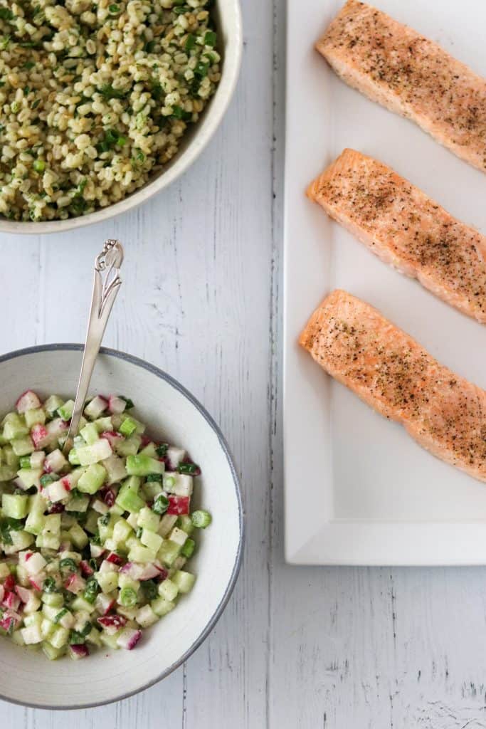 Salmon filets on a platter next to cucumber radish relish and barley salad in bowls