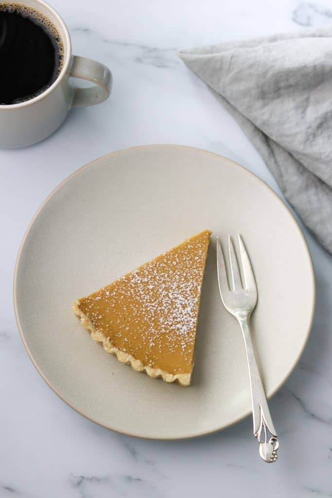 A slice of brown sugar skyr tart on a plate with a fork, napkin and a cup of coffee