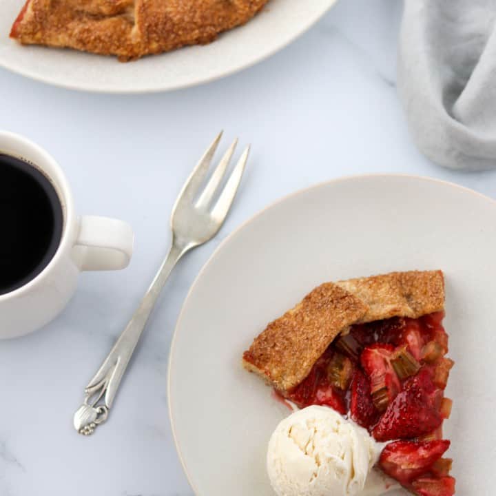 A slice of strawberry rhubarb galette with a scoop of ice cream and a fork and a cup of coffee