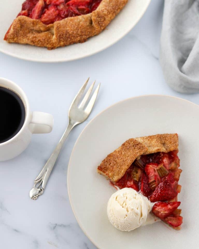 A slice of strawberry rhubarb galette with a scoop of ice cream and a fork and a cup of coffee