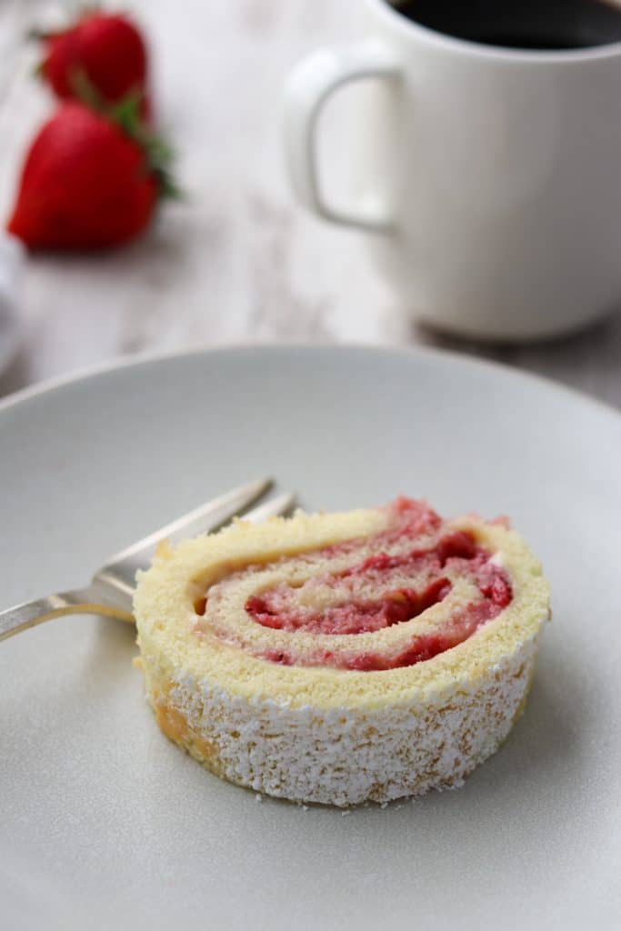 A piece of strawberry rolled cake on a plate next to a cup of coffee