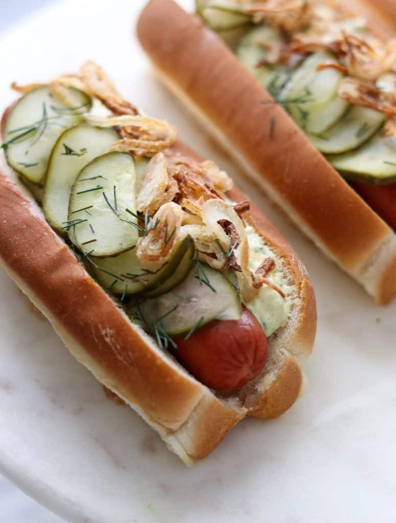 A close up of a hot dog on a bun covered in pickled cucumbers, crispy shallots and creamy sauce