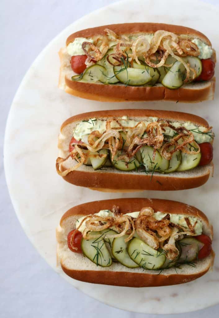 Hot dogs on buns with pickled cucumbers, crispy shallots and creamy sauce on a plate