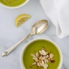 A bowl of zucchini soup topped with sliced almonds and a spoon and lemon wedge