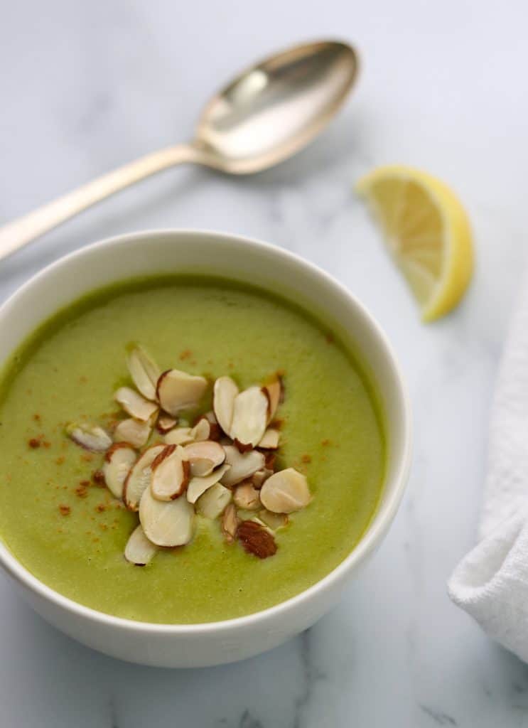 A bowl of zucchini soup topped with almonds with a lemon wedge and spoon