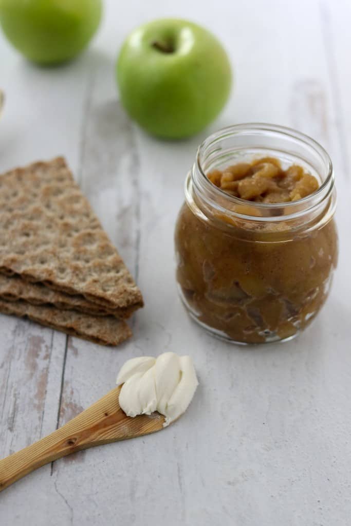 Caramelized apple compote in a jar next to apples, crispbread and a wooden knife with cream cheese