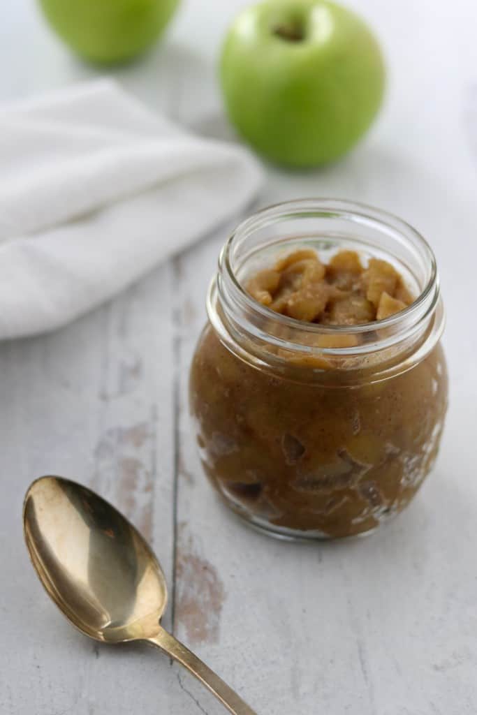 Caramelized apple compote in a jar next to a spoon, towel and apples