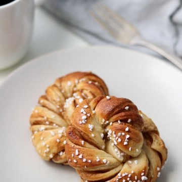 Two Swedish cinnamon buns on a plate next to a mug of coffee, fork and napkin