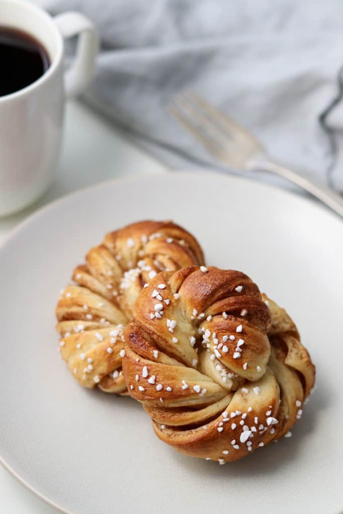 Two Swedish cinnamon buns on a plate next to a mug of coffee, fork and napkin