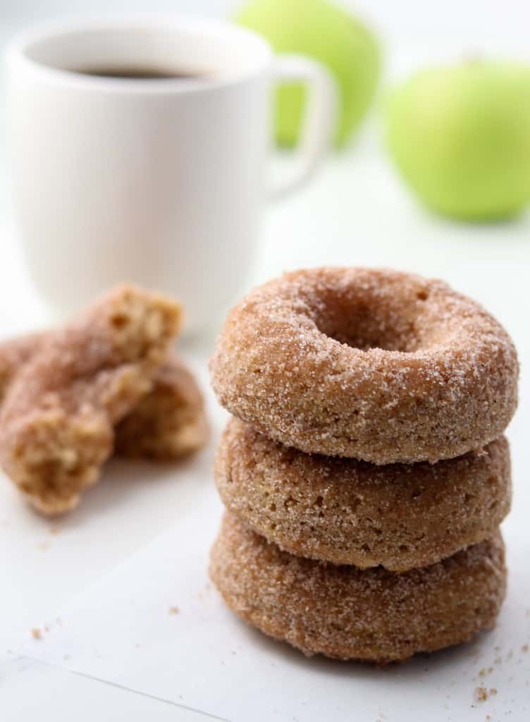 Close up of stack of three apple donuts next to a cup of coffee and apples