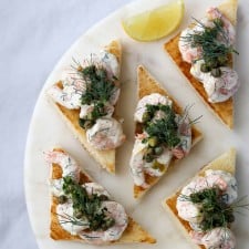 Toast triangles topped with shrimp salad, capers and dill next to lemon wedges