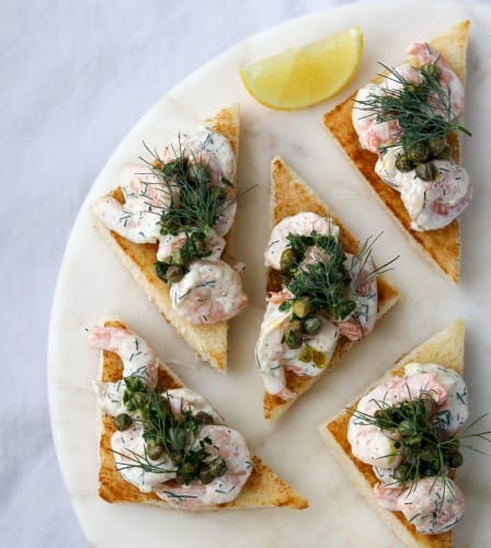 Toast triangles topped with shrimp salad, capers and dill next to lemon wedges