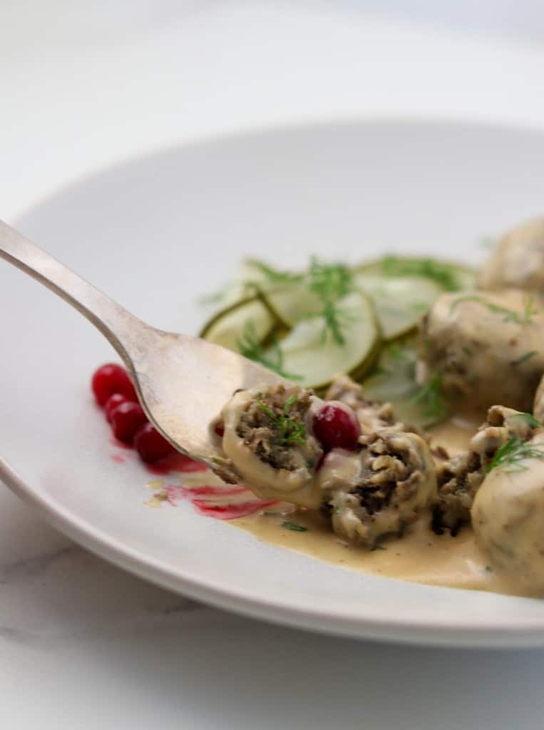 Close up of a Swedish meatball in cream sauce on a fork with lingonberries and pickles