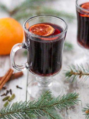 A cup of mulled wine next to evergreen branches, spices and an orange.